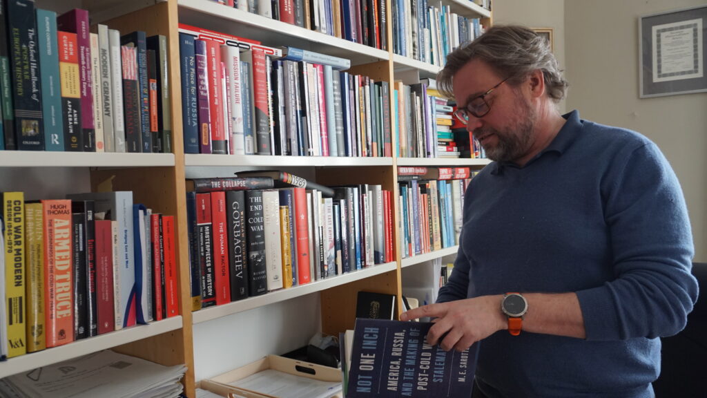 En bild på Juhana Aunesluoma. Han står framför en bokhylla med boken Not one Inch i händerna.