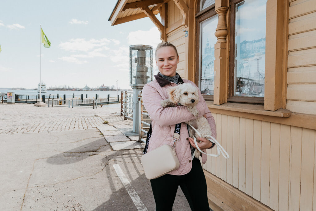 En bild på Ulrika Olsio. hon står bredvid en gul stuga med en vit hundvalp i famnen.