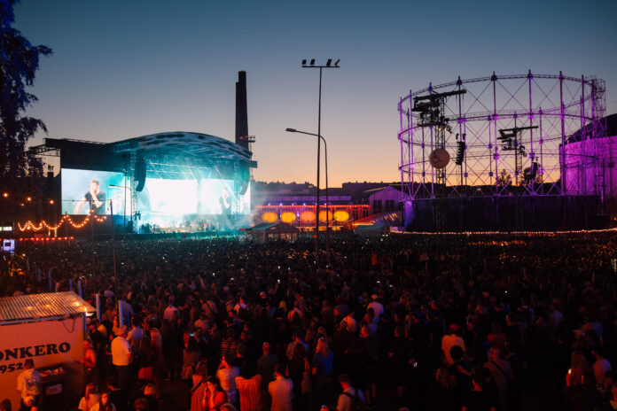 På bilden är en scene där ett musikuppträdande pågår med en stor publik framför i festival området. Det är solnedgång.