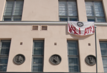 Protest mot regeringen – gymnasiestuderande marscherar ut En person går in i en rosa stenbyggnad. Ut ur ett av fönstren hänger en banderoll där det står "vallattu", alltså ockuperat.