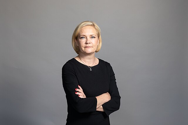 Mari Rantanen, en vit blond kvinna i 40-årsåldern med blåa ögon och kort hår, står med armarna i kors och tittar in i kameran med ett neutralt ansiktsuttryck.